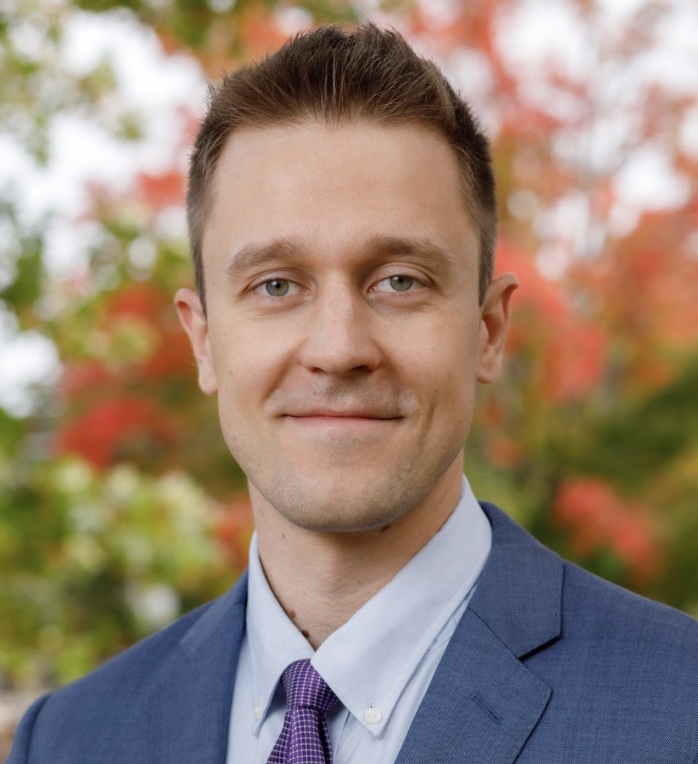 A man in a blue suit and purple tie smiles slightly, standing outdoors with blurred green and red autumn foliage in the background.