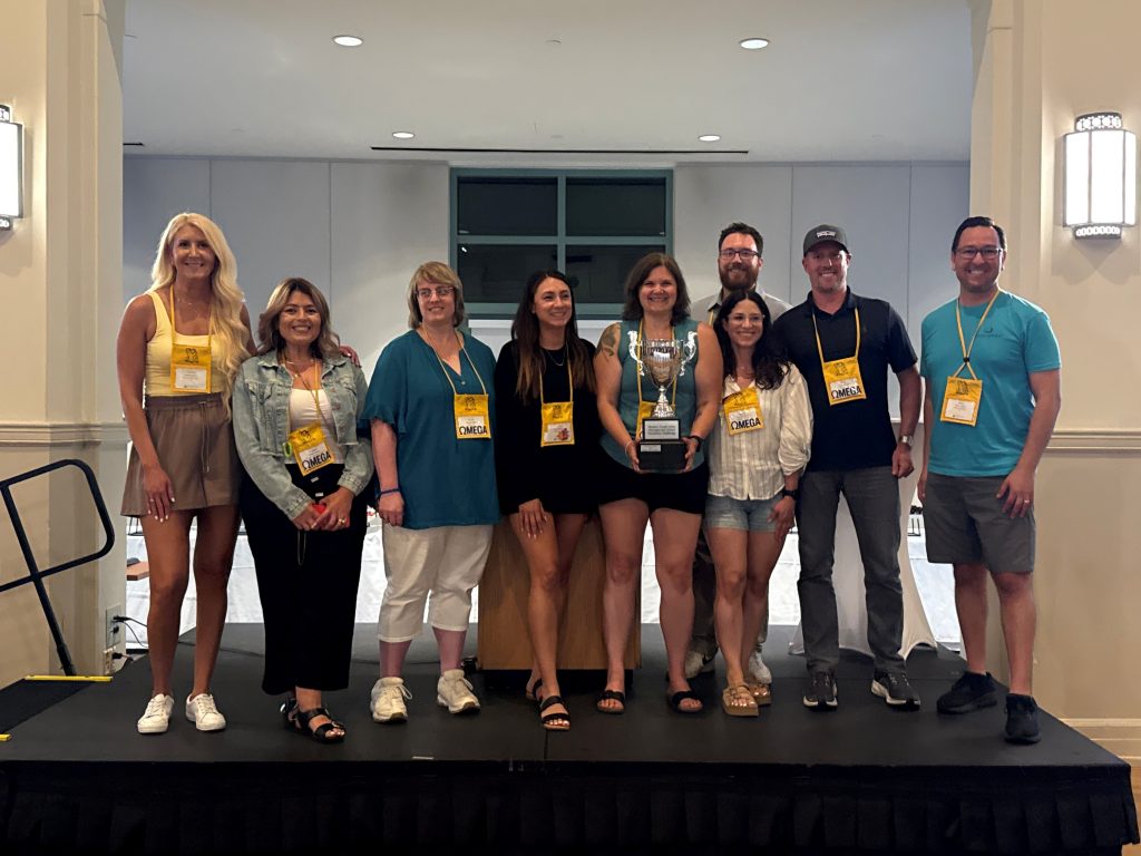 Nine people stand on a stage, smiling at the camera. One woman in the center holds a large trophy. Most wear yellow lanyards. The background is a light-colored hallway.