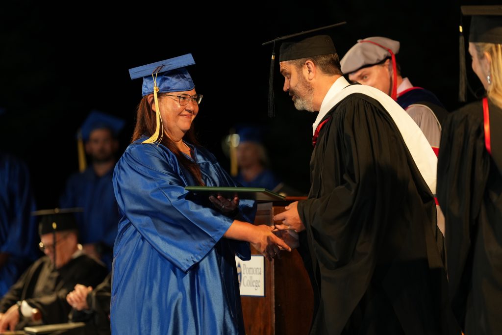 A graduate in a blue cap and gown smiles and shakes hands with a faculty member in black robes while receiving her diploma on stage during a graduation ceremony.