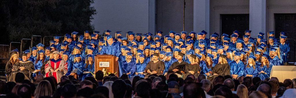 A large group of graduates in blue caps and gowns sits on stage during a commencement ceremony, with an official speaking at a podium as the audience watches.