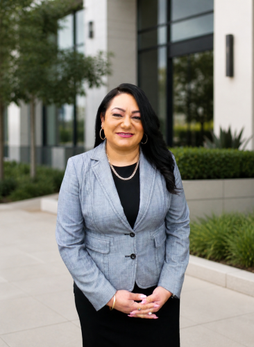 Annette Vega-Young A woman in a gray blazer and black dress stands outside a modern building, smiling at the camera. She has long dark hair and is wearing gold jewelry. Green bushes and trees are in the background.