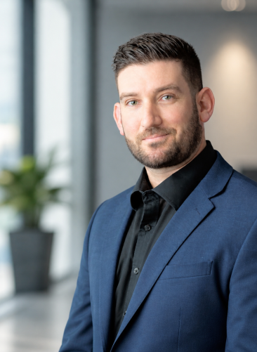 Broc Corp Background A man with short dark hair and a beard, wearing a blue suit and black shirt, stands indoors in front of large windows with a blurred plant in the background.