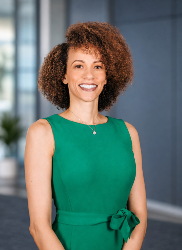 Destiny Corp Background A woman with curly hair and a green sleeveless dress smiles while standing in a modern, blurred indoor setting.
