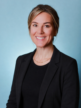 A woman with blonde hair in a low updo, wearing a black blazer over a black top, and a gold necklace, smiles in front of a light blue background.