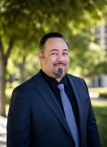George Zoomed Out A man with dark hair and a goatee, wearing a dark suit, black shirt, and patterned tie, stands outdoors with trees and greenery in the background.