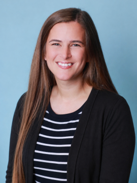 A woman with long brown hair, wearing a black cardigan over a black and white striped shirt, smiles in front of a light blue background.