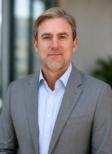 Ian Wingfield A man with light hair and a beard, wearing a gray suit and blue striped shirt, stands outside in front of a blurred building and greenery.